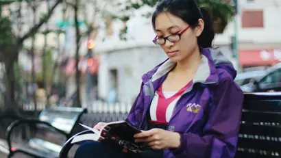 A woman in glasses and a purple jacket is sitting on a park bench reading a book