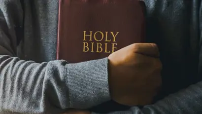 A person wearing a gray sweater holding a maroon colored Bible