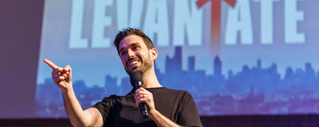 A smiling man in a black shirt holds a microphone while pointing away from himself.