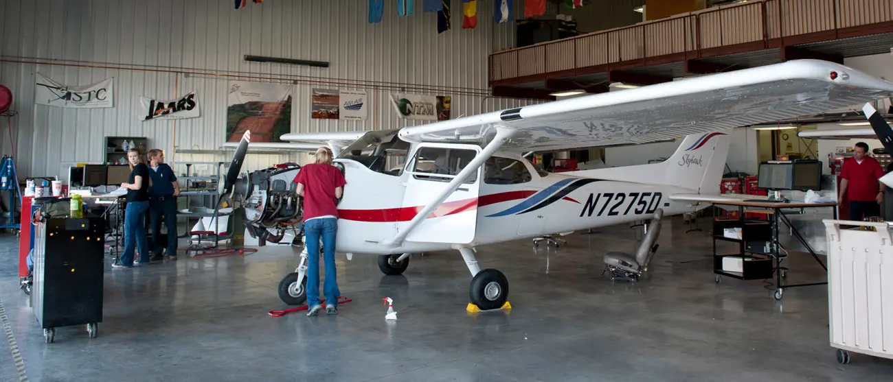 A man in a red shirt works on a small plane in a hangar. 